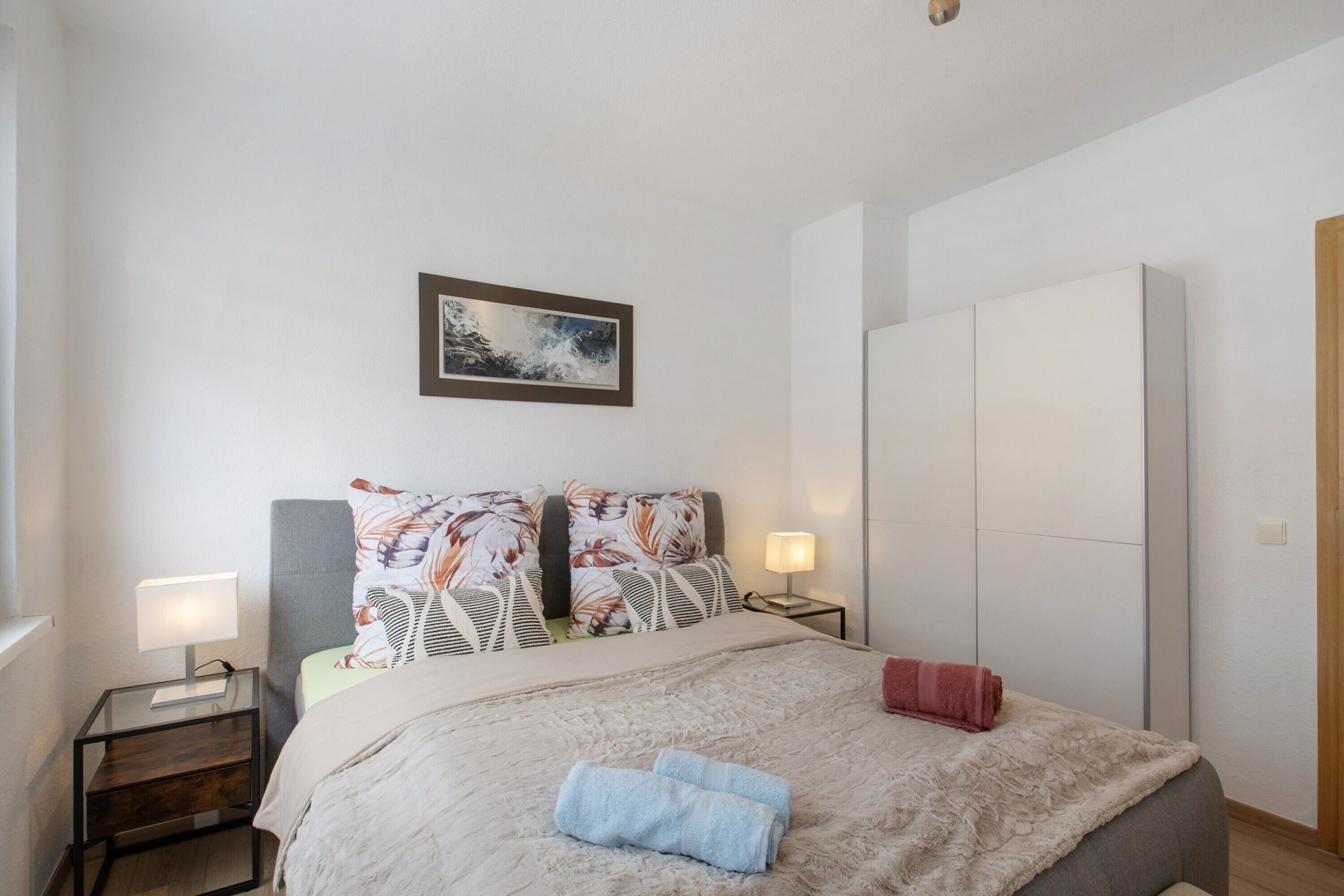 Quarto com cama, duas mesas de cabeceira, um criado-mudo, uma obra de arte na parede e um guarda-roupa.