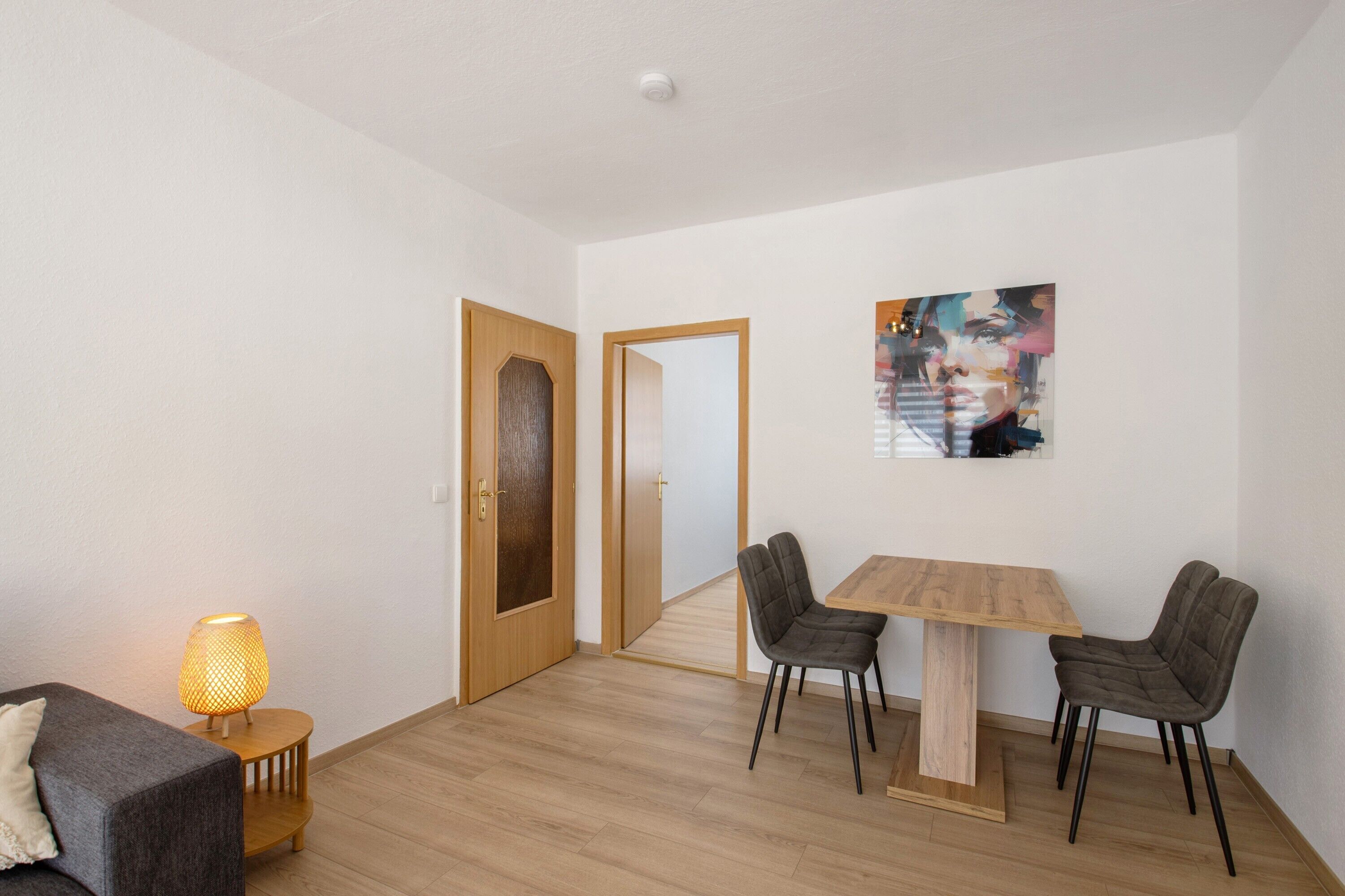 A room with a wooden floor, a dining table with four chairs, a lamp, and a painting on the wall.