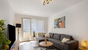 A modern living room with a sofa, coffee table, and lamp. There is a TV, a plant, and a painting on the wall.