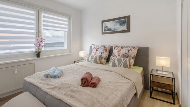 A bedroom with a bed, two bedside lamps, a window with blinds, a potted plant, and a painting on the wall.