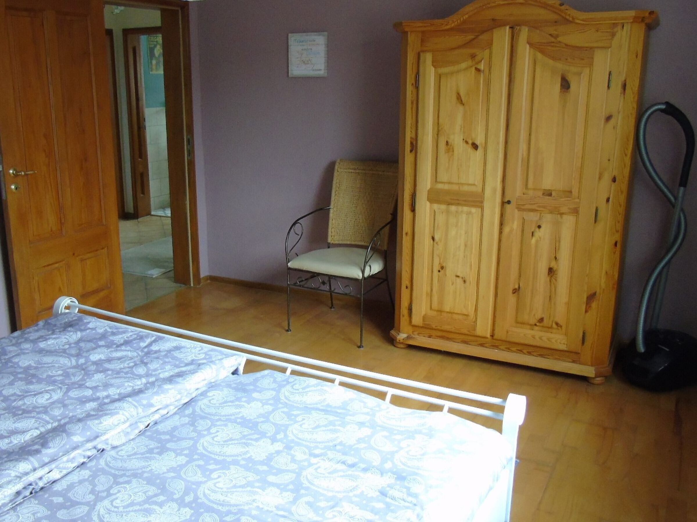 A bedroom with a bed, a wooden wardrobe, a chair, and a vacuum cleaner.