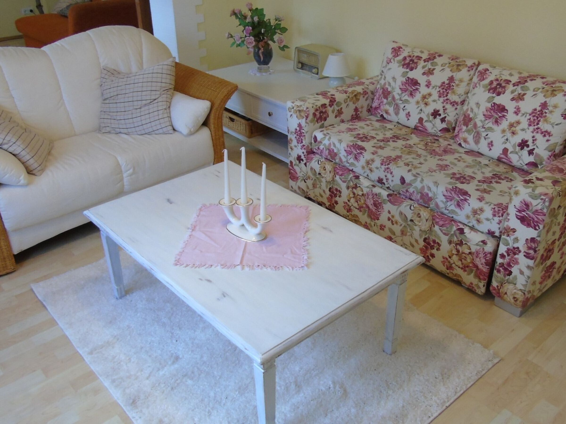Ein Wohnzimmer mit einem weißen Sofa, einem blumigen Sofa, einem weißen Couchtisch mit Kerzen und einem Radio auf einem Beistelltisch.