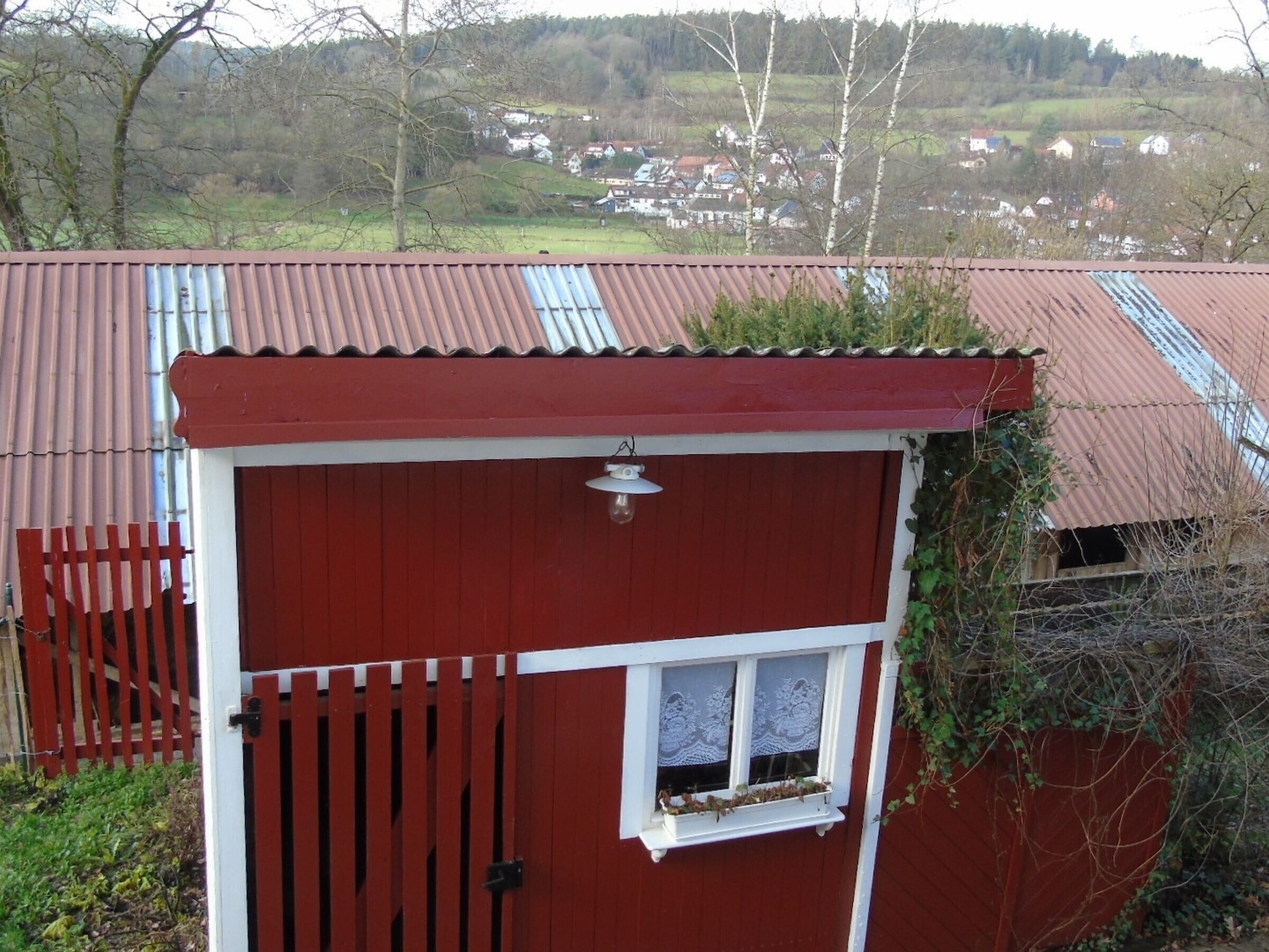 Una estructura de madera roja con techo de láminas metálicas, una pequeña ventana con cortinas de encaje y una puerta blanca.