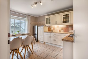 Una cocina con alacenas blancas, un refrigerador, una mesa de comedor con sillas y una ventana con cortinas.