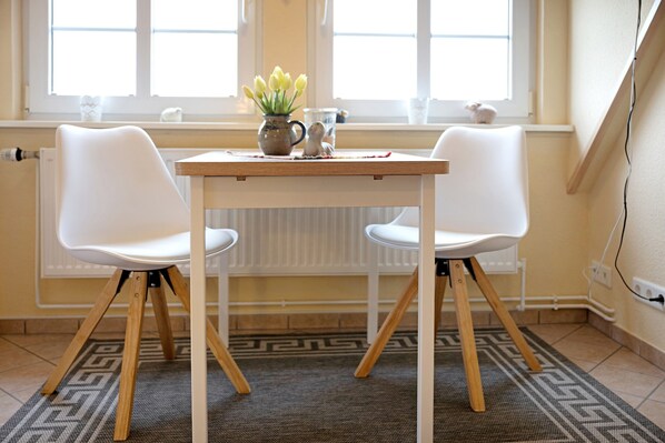 Une pièce avec une table et deux chaises, une fenêtre donnant sur un paysage agréable, un radiateur et un tapis à motifs.