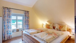 Een slaapkamer met een houten bed, bloemenbeddengoed, een raam met gordijnen en een nachtkastje met een lamp.