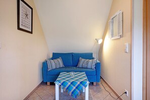 Une pièce avec un canapé bleu, des coussins à rayures, une nappe à carreaux, une horloge murale et une fenêtre avec des volets.