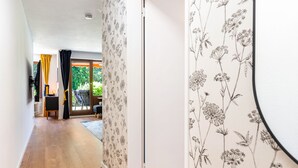 A hallway with wooden flooring, a door with floral wallpaper, and a view of a balcony with yellow curtains.