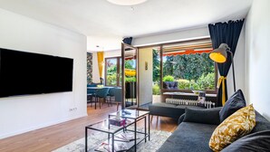 A modern living room with a flat-screen TV, a sofa, a coffee table, and a view of a garden through large windows.