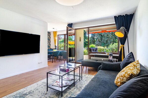 A modern living room with a flat-screen TV, a sofa, a coffee table, and a view of a garden through large windows.