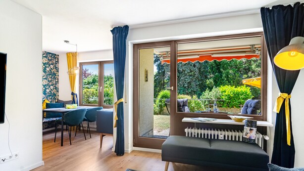 A modern living room with a sofa, a dining area with chairs, and a large sliding glass door leading to a garden.
