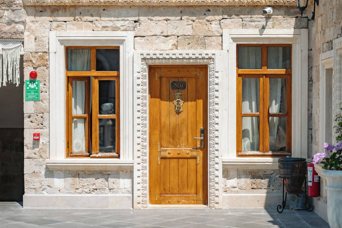 Edificio de piedra con puerta de madera marcada '301', dos ventanas con cortinas, un extintor y un letrero que indica una salida de emergencia en el último piso.