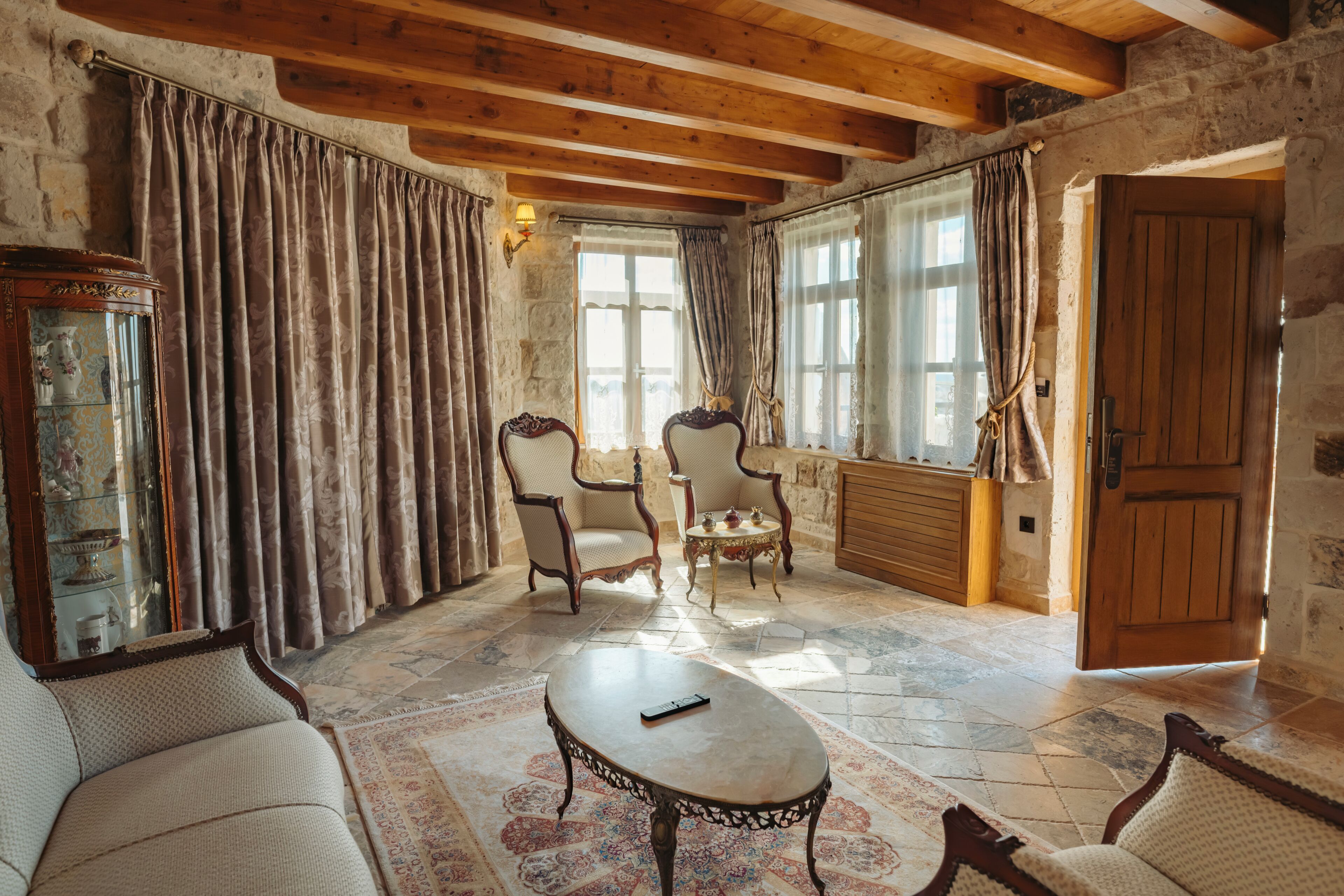 A rustic room with stone walls, wooden beams, a fireplace, two armchairs, a coffee table, and a wooden door.