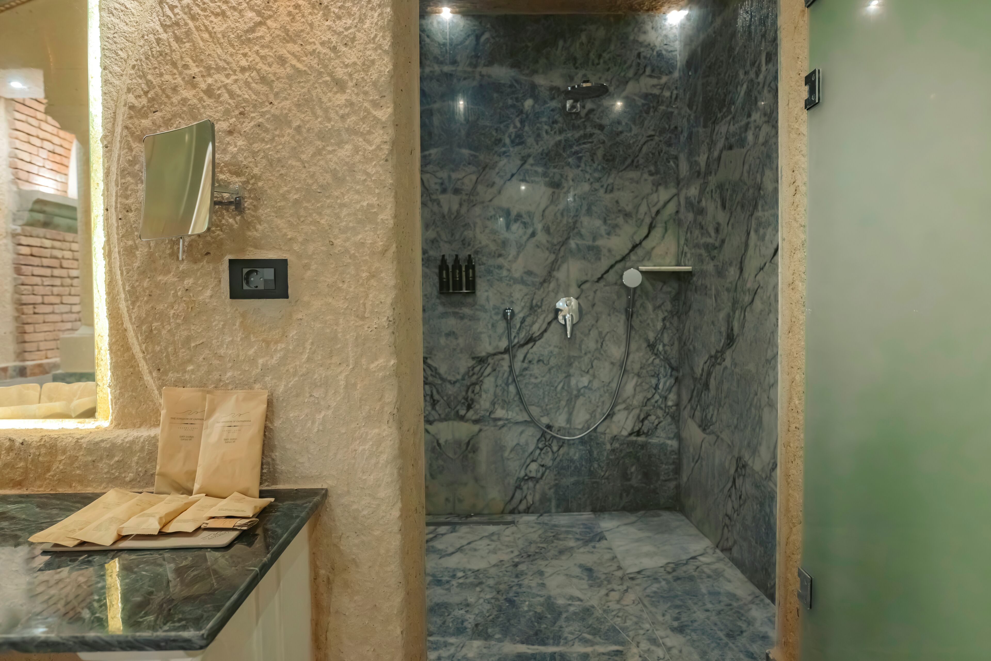 A bathroom with a marble shower, a sink with a mirror, and a window with a view of a brick wall.