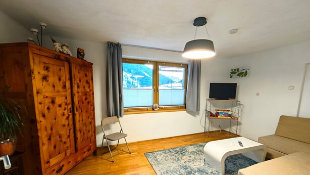 Ett vardagsrum med en trägarderob, en soffa, en tv på ett tv-bord, ett fönster med utsikt över bergen och en hängande lampa.