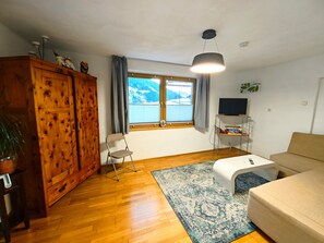 A living room with a wooden wardrobe, a sofa, a TV on a stand, a window with a view of mountains, and a hanging light fixture. - Apartment 'Mizi' with Mountain View, Balcony and Wi-Fi (Stall)