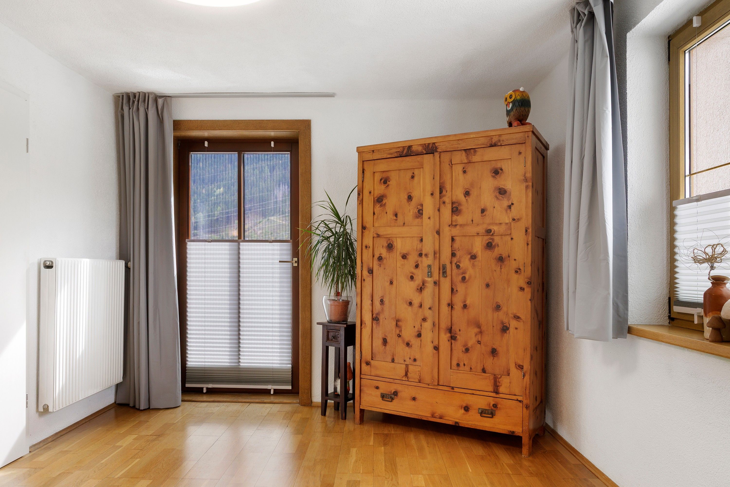Ein Zimmer mit einem Holzschrank, einem Heizkörper, einer Zimmerpflanze und einem Fenster mit Vorhängen.
