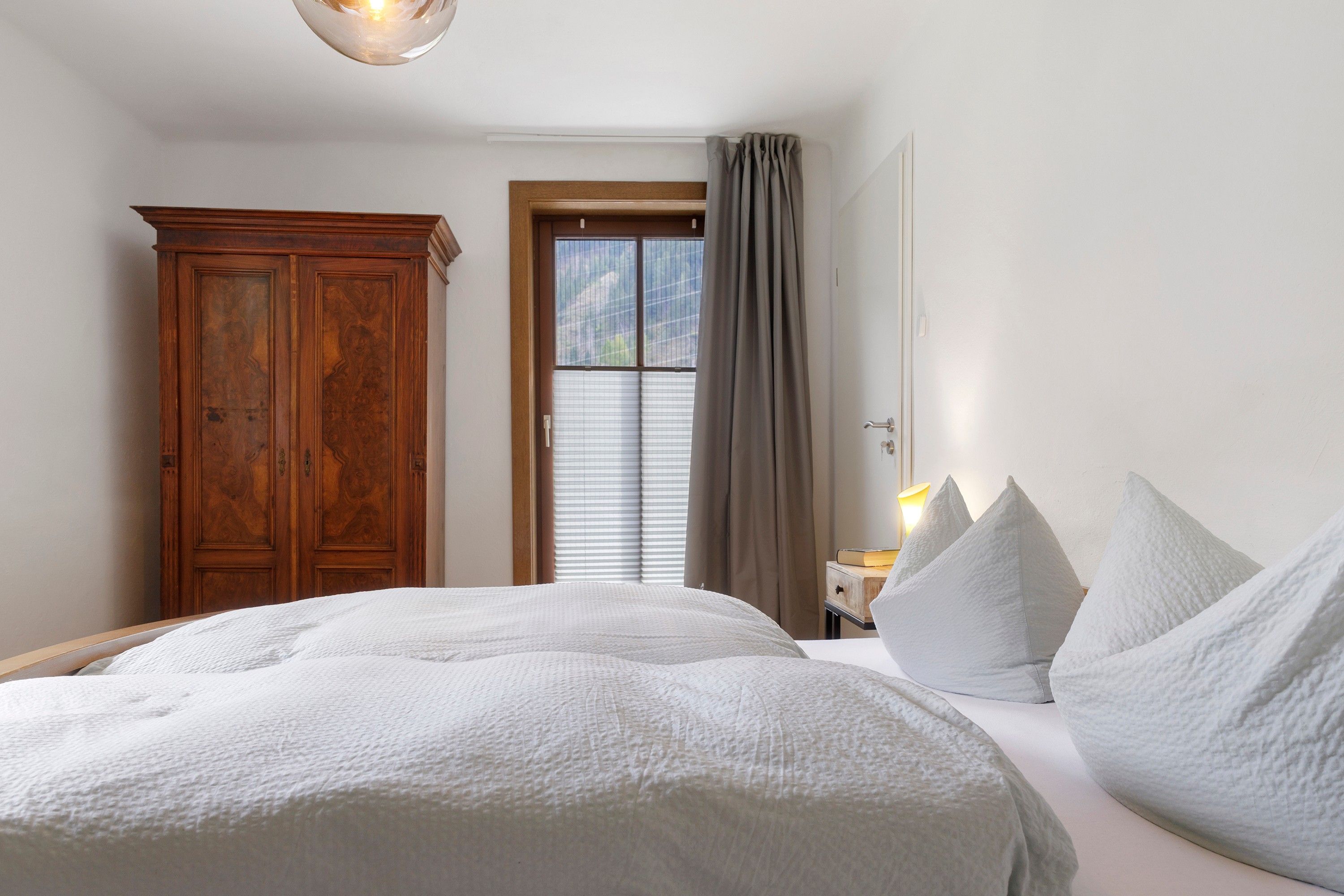 A bedroom with a large bed, a wooden wardrobe, a window with curtains, and a bedside table with a lamp.