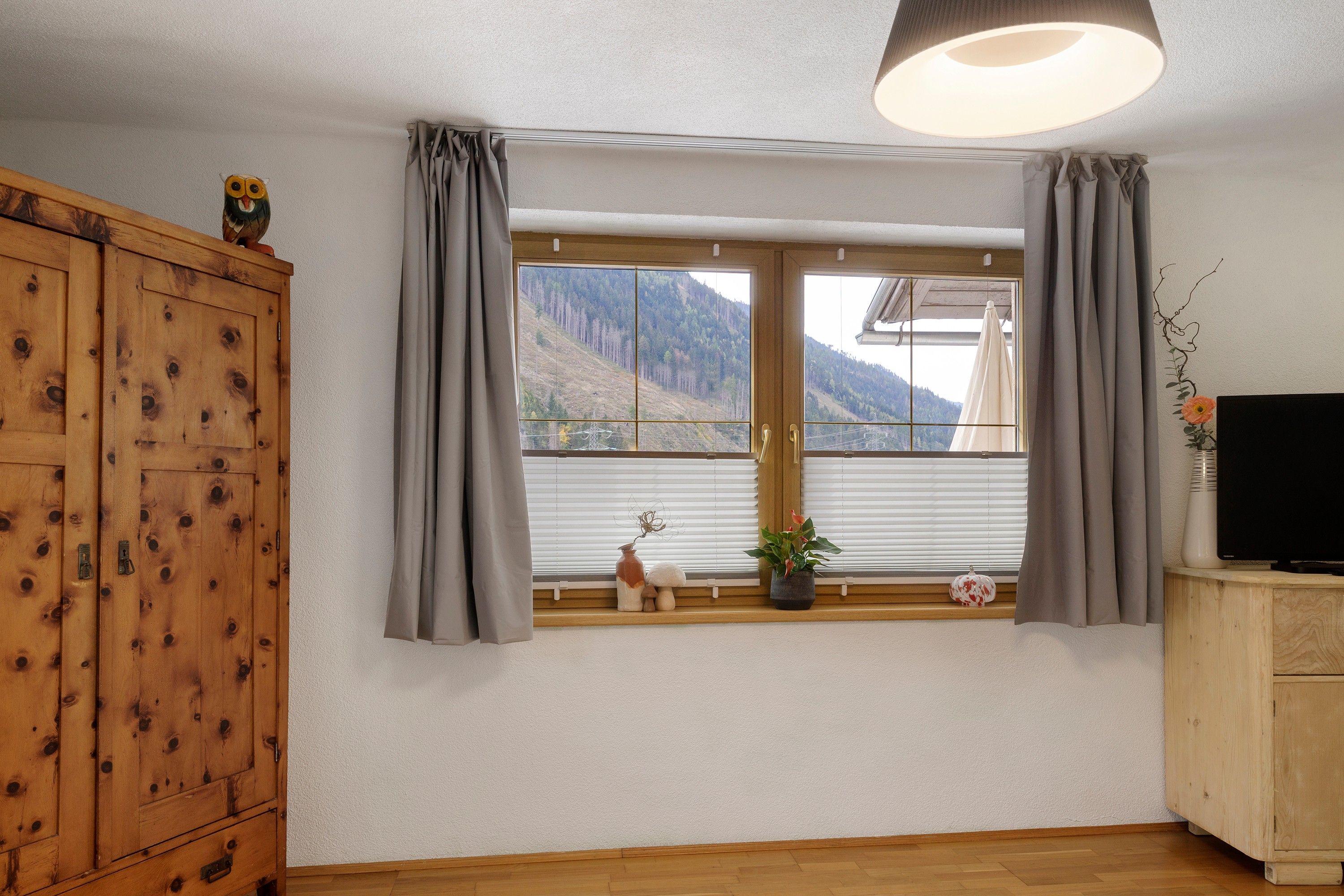 A room with a large window, wooden wardrobe, and a television on a wooden cabinet.