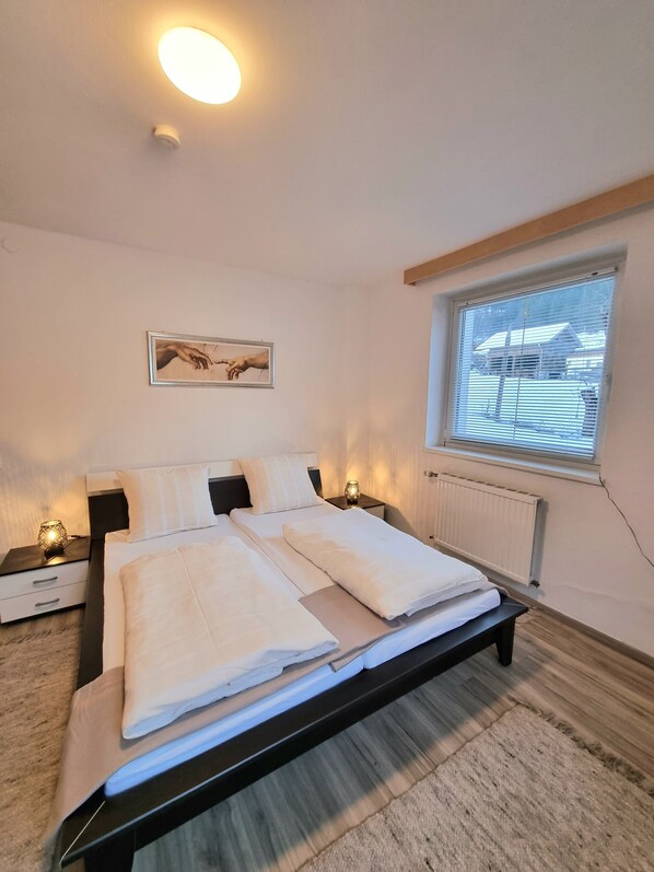 A bedroom with a bed, bedside tables, a window with blinds, a radiator, and a painting on the wall.