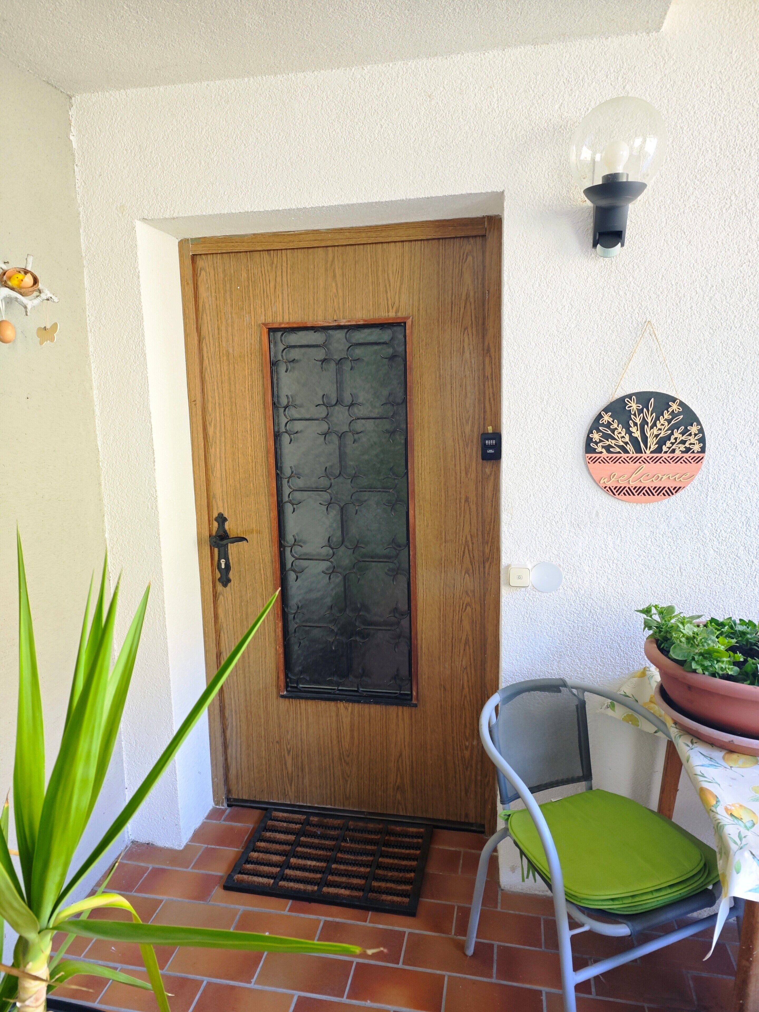 Uma porta de madeira com um painel de vidro decorativo, uma cadeira de metal com almofada verde, uma planta em vaso e um aplique de parede.
