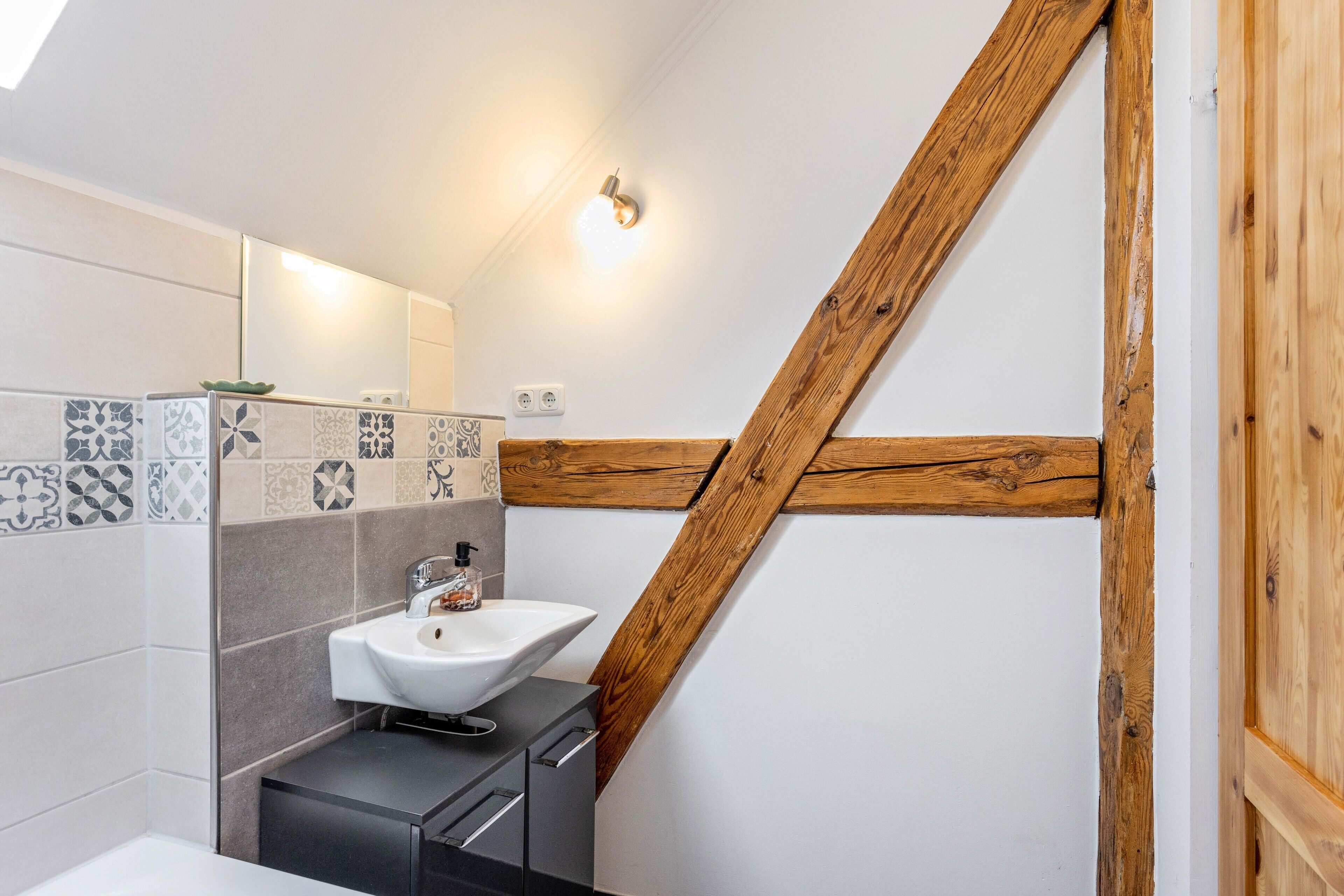 Ein Badezimmer mit weißem Waschbecken, gefliesten Wänden, einem Holzbalken und einer Leuchte.