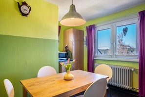 A dining area with a wooden table, white chairs, a vase with flowers, a wall clock, and a window with curtains.