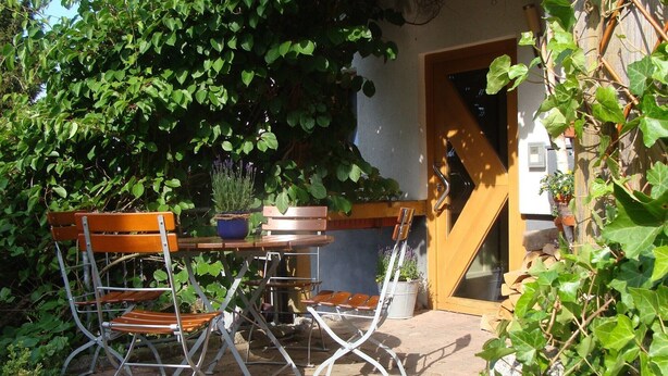 An outdoor patio with a wooden table and metal chairs, surrounded by greenery and a wooden door. - Apartment 'Blackforest' with Shared Terrace, Shared Garden and Wi-Fi (Sankt Georgen)