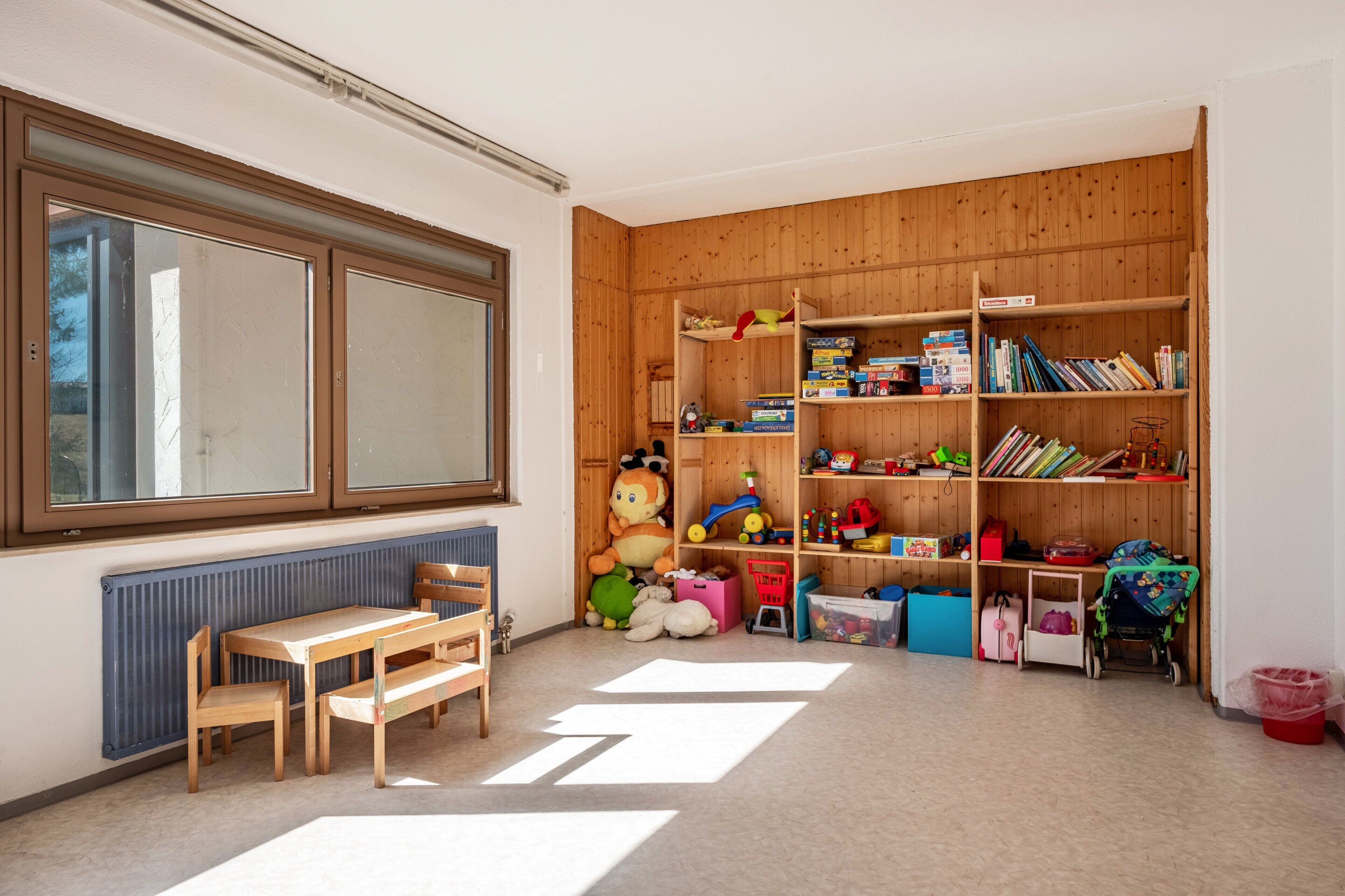Ein Zimmer mit einem Bücherregal aus Holz, einem Fenster, einer Heizung und einem Tisch mit Stühlen.