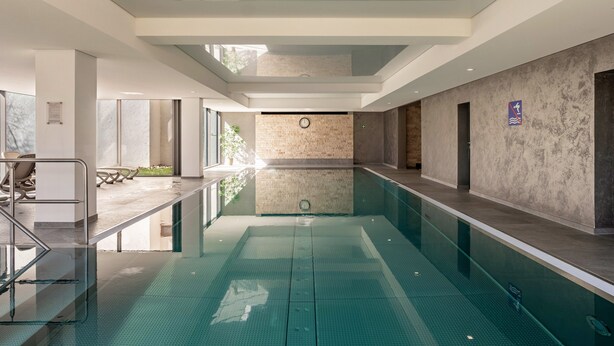 A modern indoor swimming pool with clear glass walls, a tiled floor, and a minimalist design.