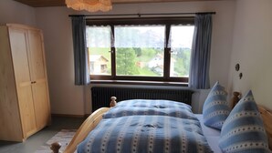 A bedroom with a wooden bed, a window with curtains, a radiator, and a view of houses outside.