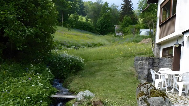 A rustic house with a stone wall, a small stream, and a garden with white plastic chairs.