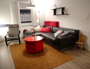 Een woonkamer met een grijze fauteuil, een rode salontafel, een zwarte leren bank met rode en grijze kussens, een houten stoel en een klein plankje met vazen.