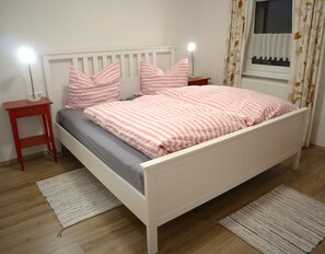 Un letto con testiera e pediera bianche, due comodini con lampade e una finestra con tende.
