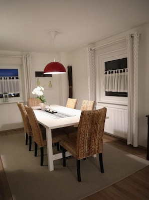 Een eetruimte met een witte tafel, rieten stoelen, een rode hanglamp en een raam met witte gordijnen.
