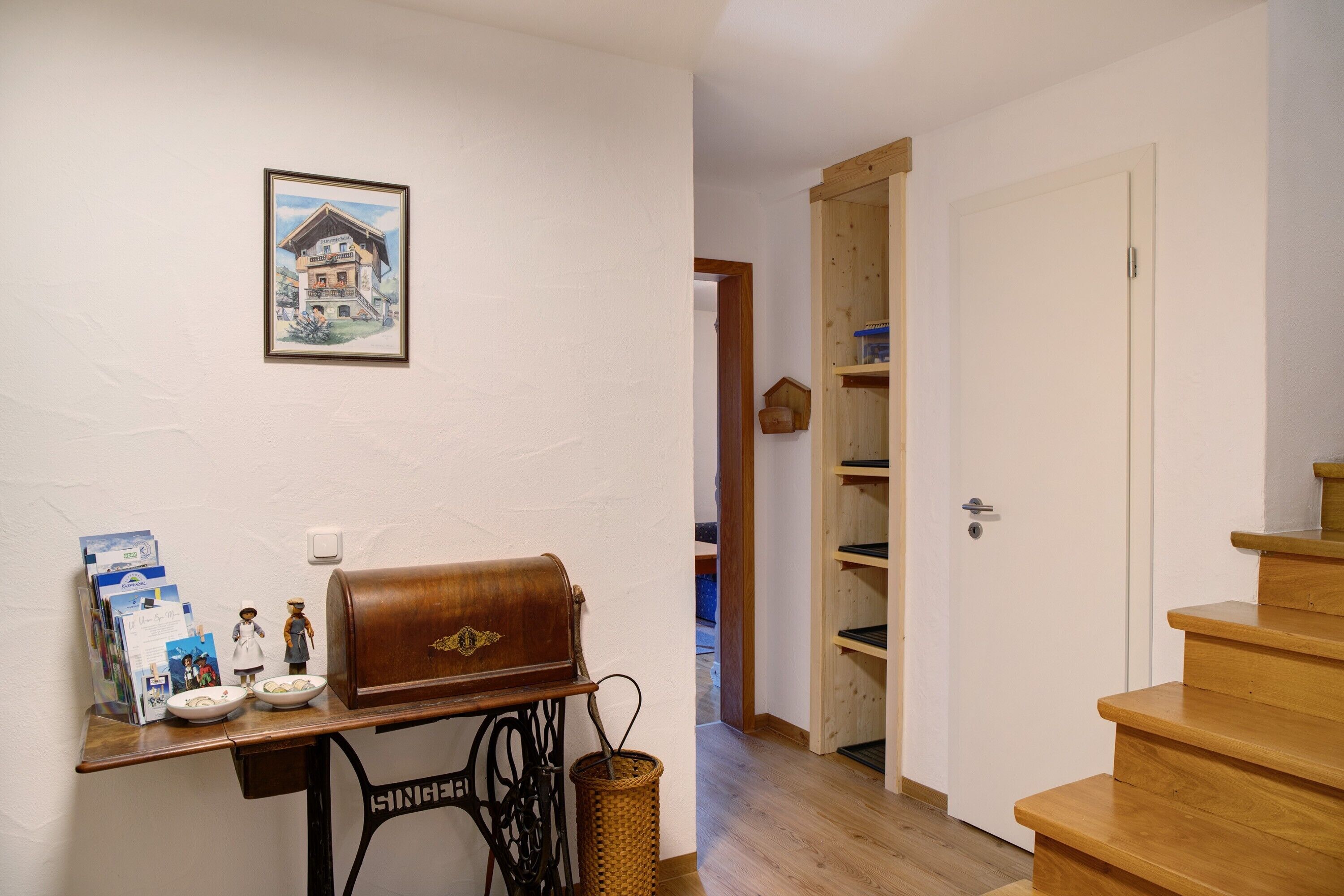 A room with a sewing machine, a wooden table with a leather bag, a framed picture, and a staircase.