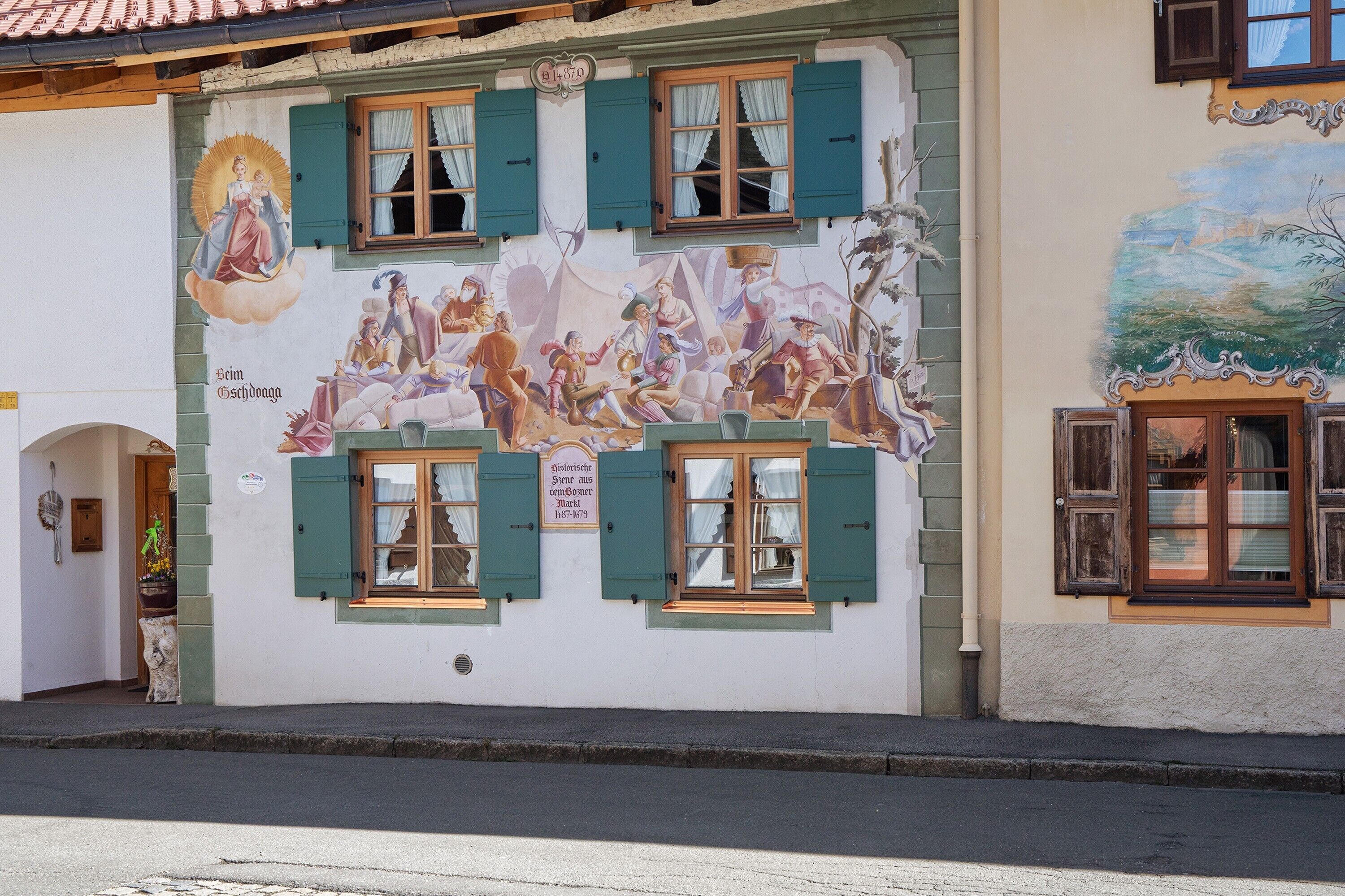 A building with a mural depicting historical scenes, green shutters, and a sign indicating a local business.