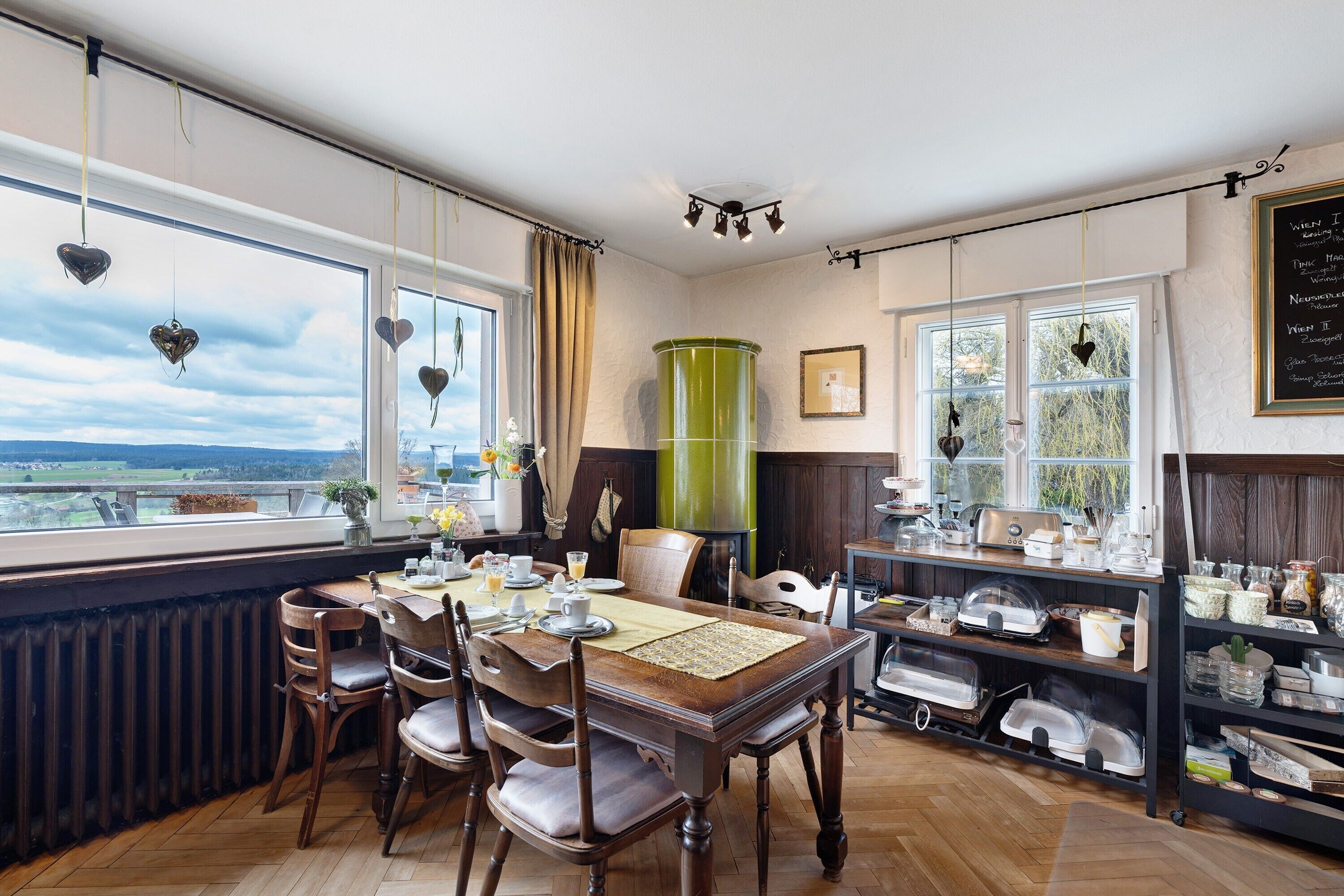 A dining area with a view, wooden chairs, a table with placemats, and a shelf with kitchenware.