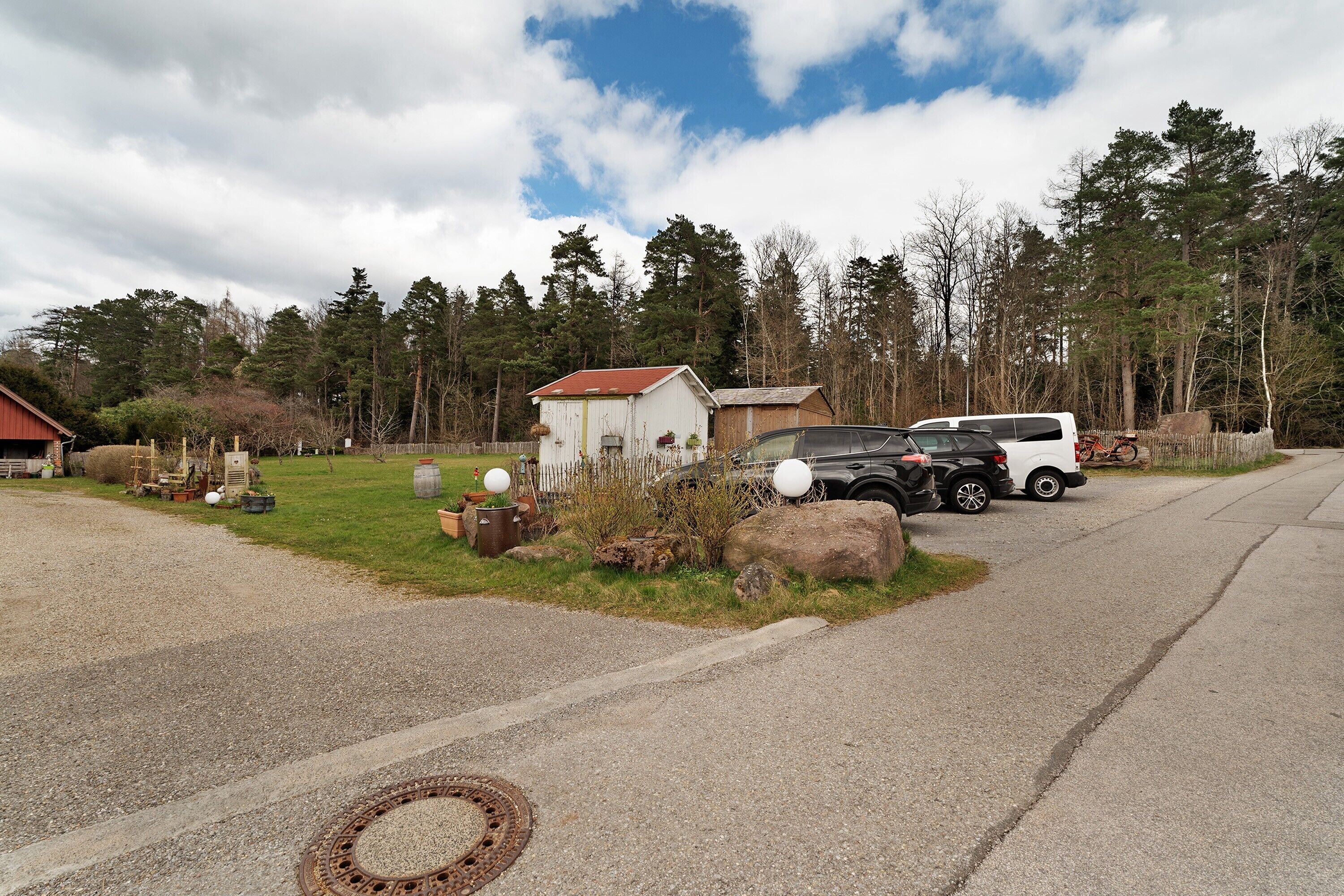 A gravel driveway leading to a white building with a red roof, parked cars, and a forested background.