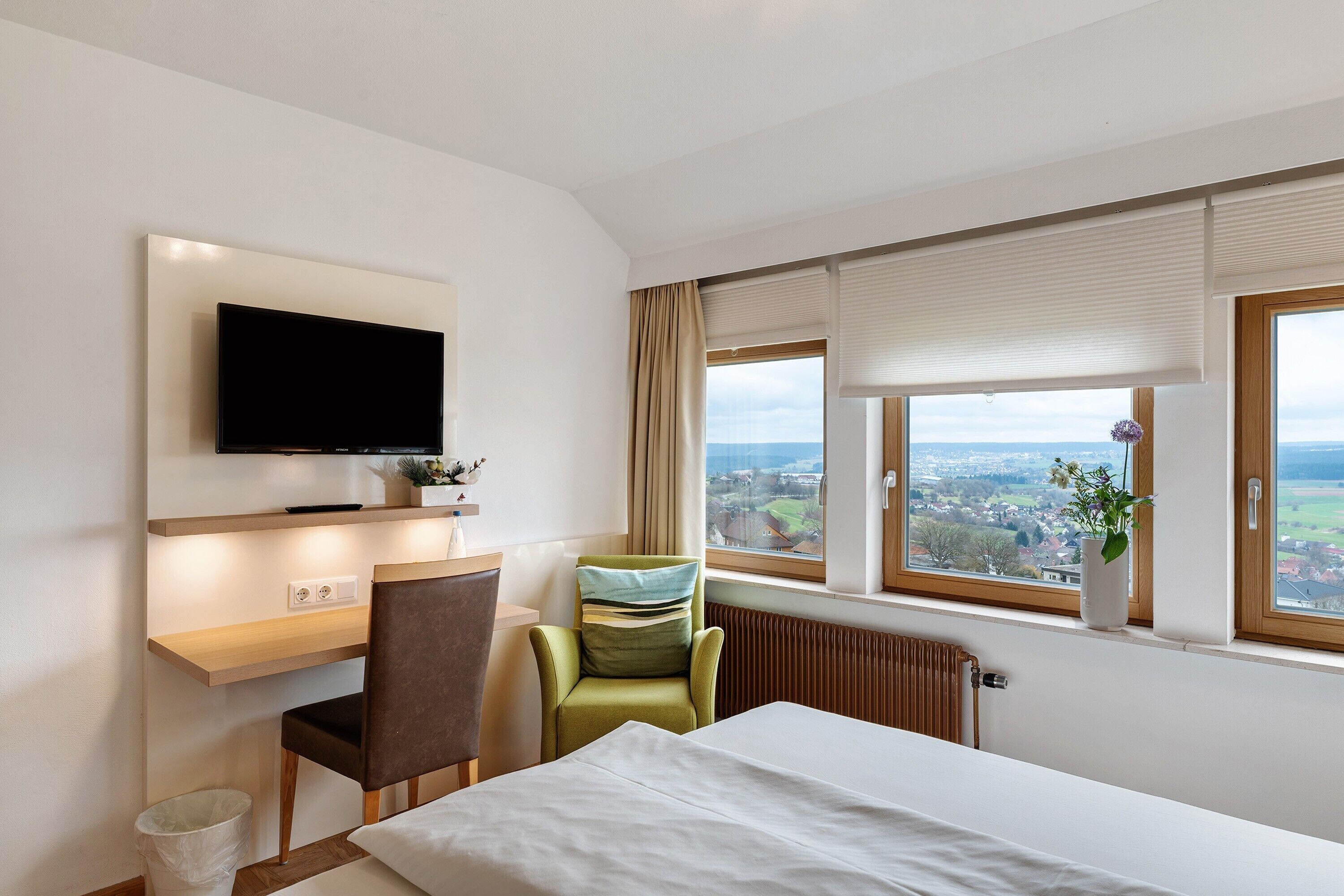 Una camera d'albergo con un letto, una scrivania con una sedia, una TV a parete, una finestra con vista sulla città e un termosifone.