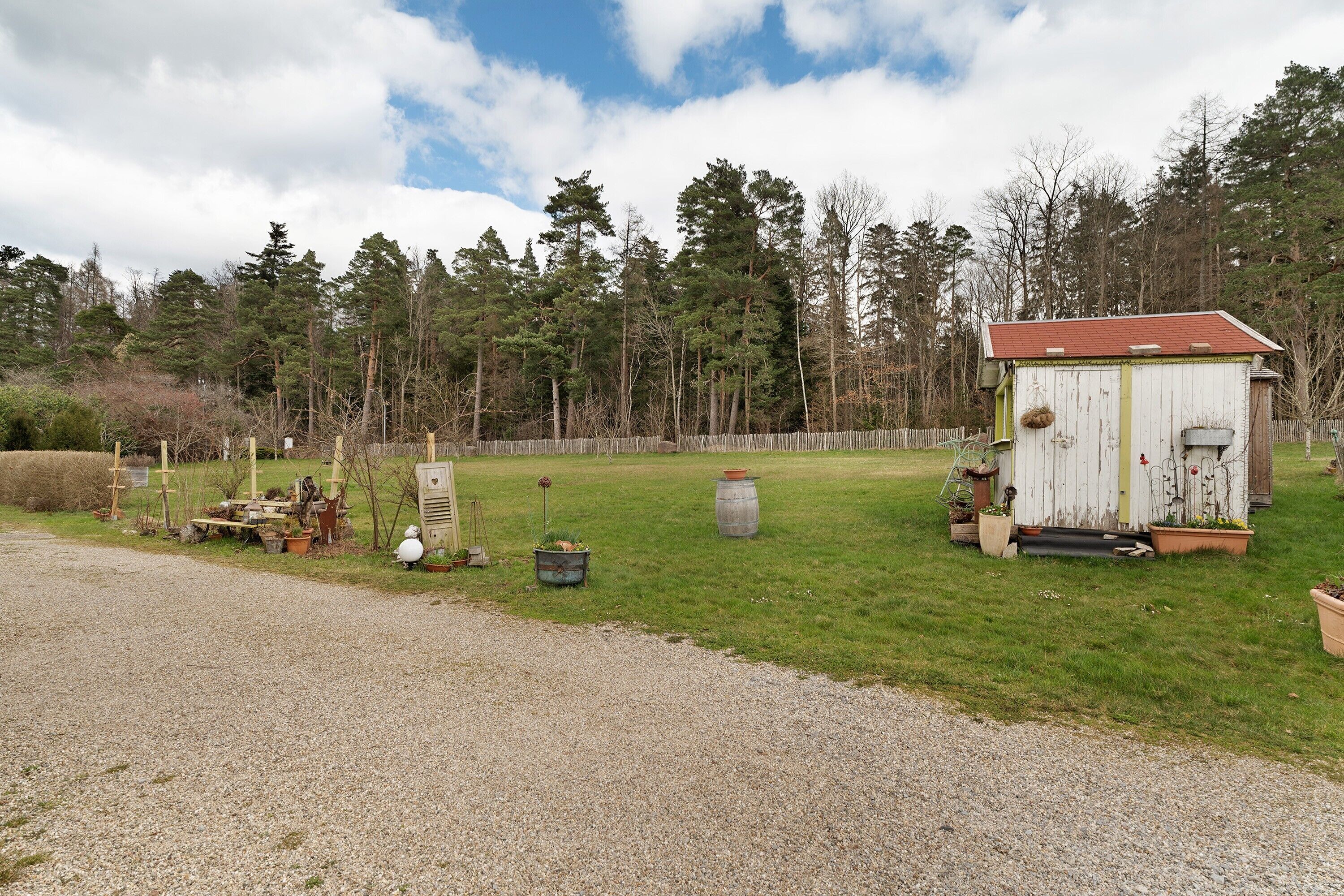 A rural property with a gravel driveway, a white shed with a red roof, and a forested background.