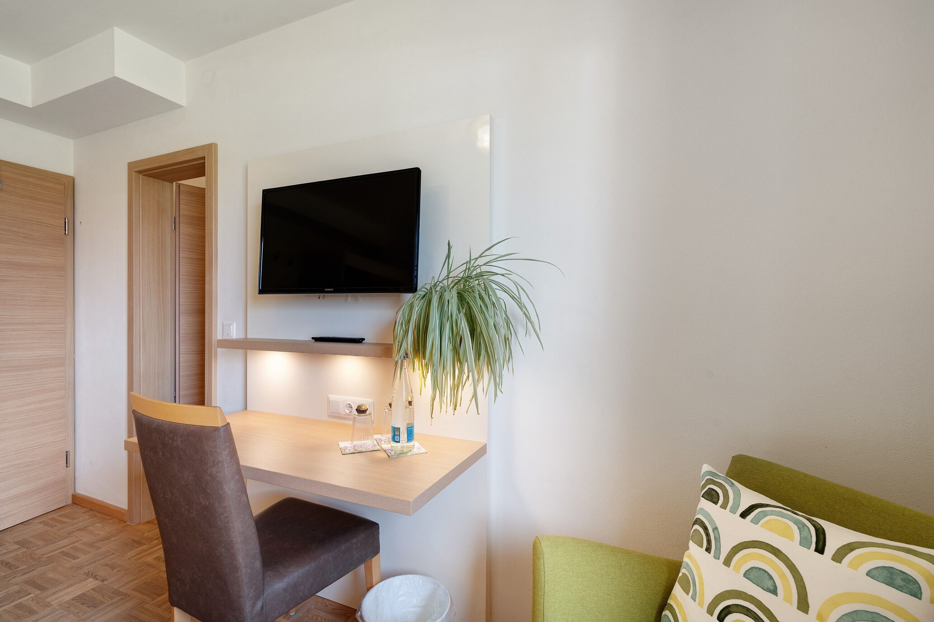 Una habitación moderna con un escritorio de madera, un televisor montado encima, una silla, una mesita pequeña y un sofá verde.