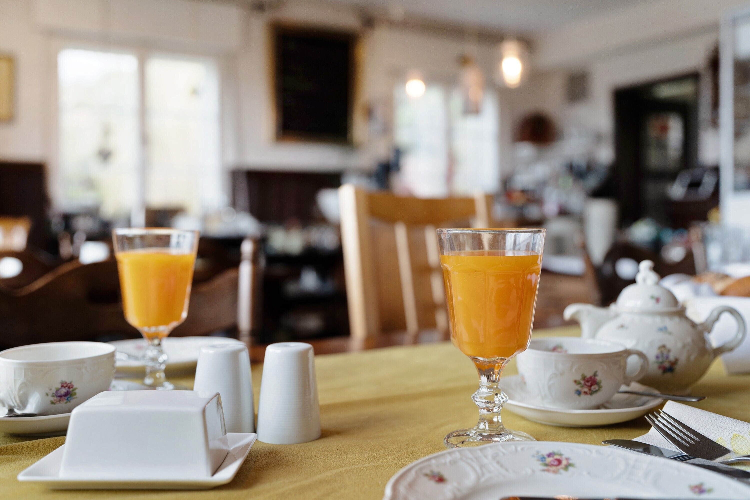 A breakfast setting with a table set for two, including a teapot, cups, and a glass of orange juice.