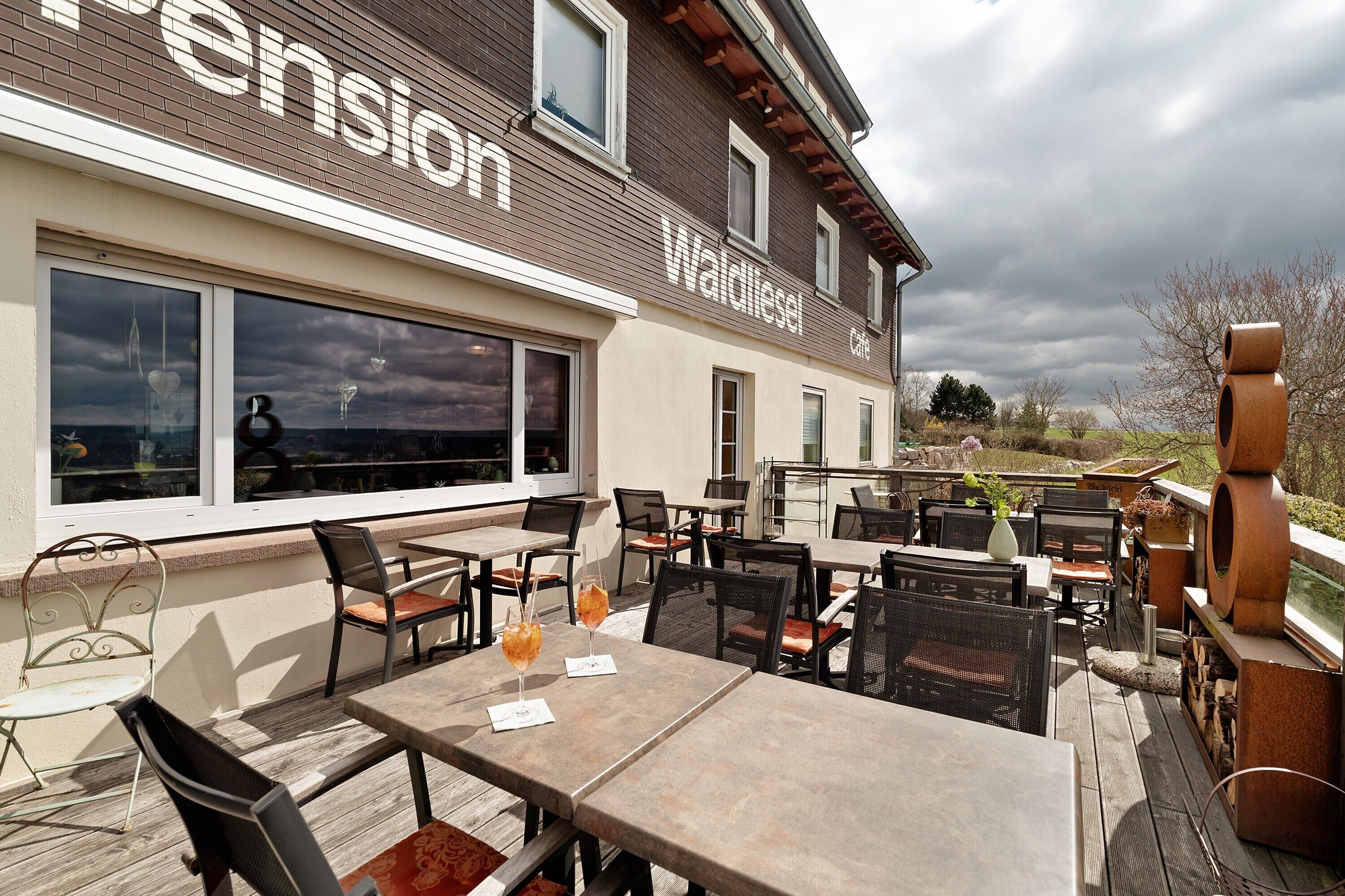Eine Terrassengastronomie mit Tischen und Stühlen, ein großes Fenster und Blick auf die Landschaft.