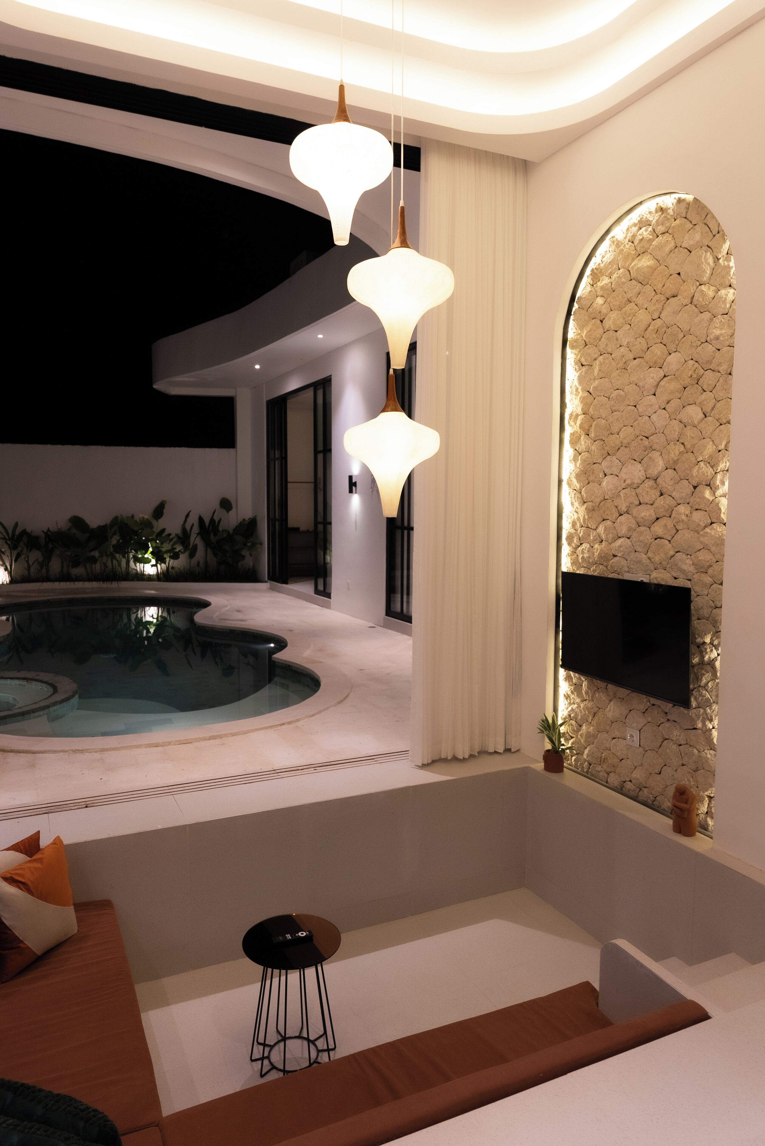 Un área de piscina interior moderna con pared de piedra, televisor de pantalla plana y lámparas colgantes.