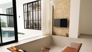 A modern living room with a large window, a flat-screen TV mounted on a textured wall, a small round table, and a sofa with cushions. - Tropical 2BR Private Villa, 6min Pererenan Beach (Munggu)