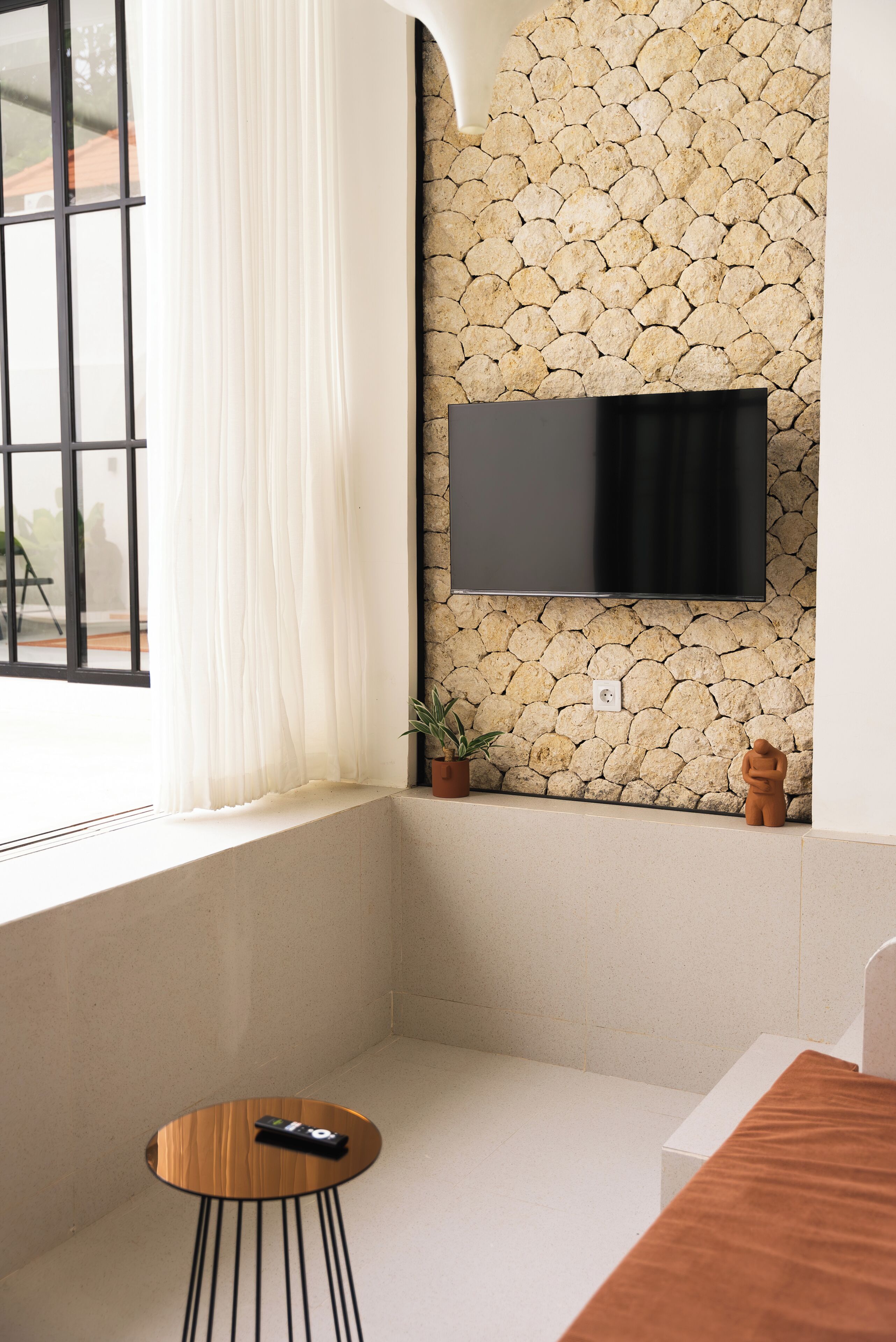 A corner room with a stone wall, a flat-screen TV mounted above a window with white curtains, a small round table with a remote control, and a potted plant.