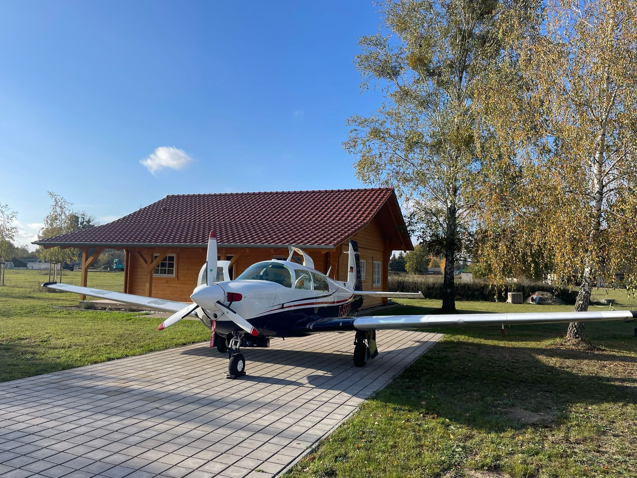 A small white and blue airplane parked on a paved area in front of a wooden cabin with a red roof.