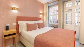 Un dormitorio con cama, mesita de noche, luz de pared y ventana con vistas a edificios.
