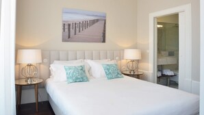 Una camera d'albergo con un letto grande, due comodini con lampade, un quadro di un molo e un bagno visibile attraverso una porta aperta.
